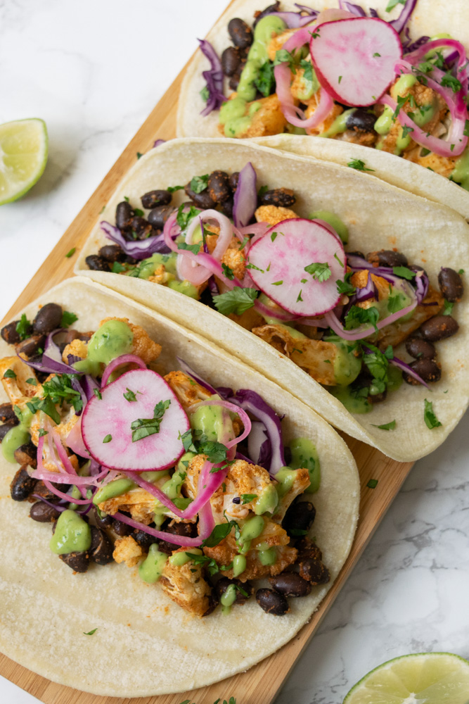 A row of cauliflower and black bean tacos topped with pickled red onions, red cabbage, radish and avocado lime crema.