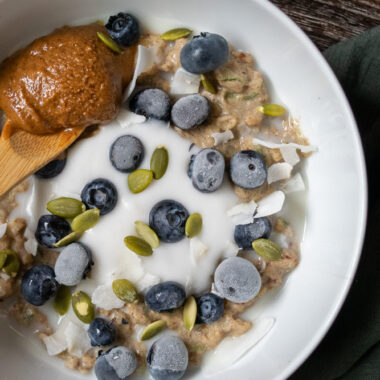 A bowl of zucchini oatmeal topped with yogurt, frozen blueberries, pumpkin seeds and a spoonful of almond butter.