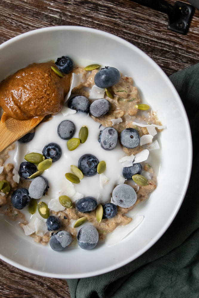 A bowl of zucchini oatmeal topped with yogurt, frozen blueberries, pumpkin seeds and a spoonful of almond butter.