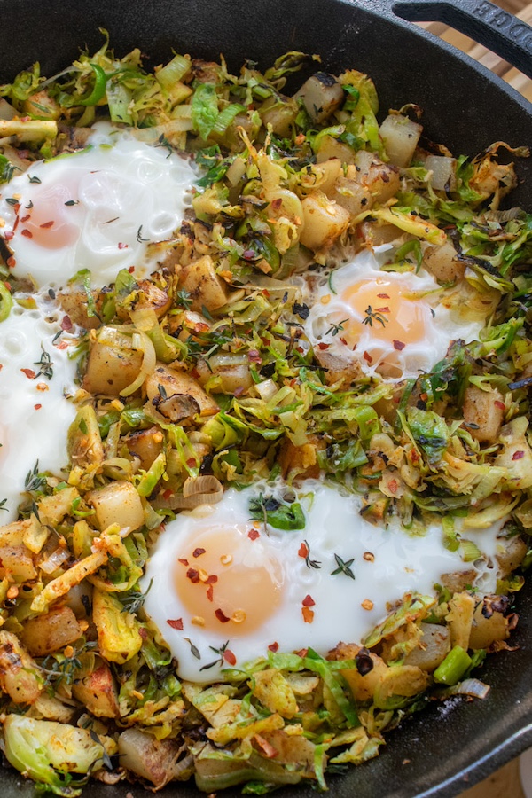 A cast iron skillet filled with a Brussels sprout and potato hash topped with cooked eggs.