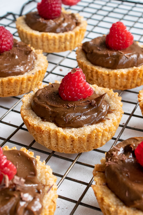 chocolate avocado mousse tartlets topped with fresh raspberries on a cooling rack.