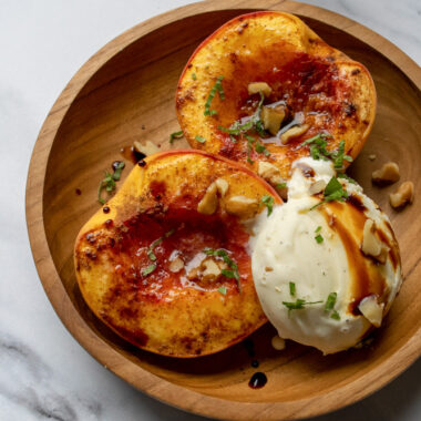 a wooden bowl filled with baked peaches with a scoop of vanilla ice cream topped with a thick balsamic vinegar, walnuts and fresh mint