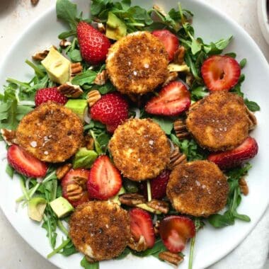 fried goat cheese and strawberry salad in a white bowl in front of sliced strawberries and a bowl of pecans.