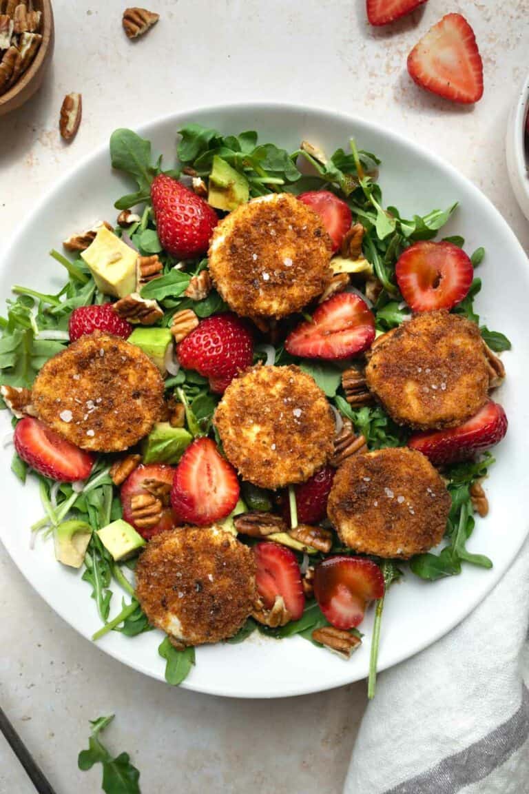 fried goat cheese and strawberry salad in a white bowl in front of sliced strawberries and a bowl of pecans.