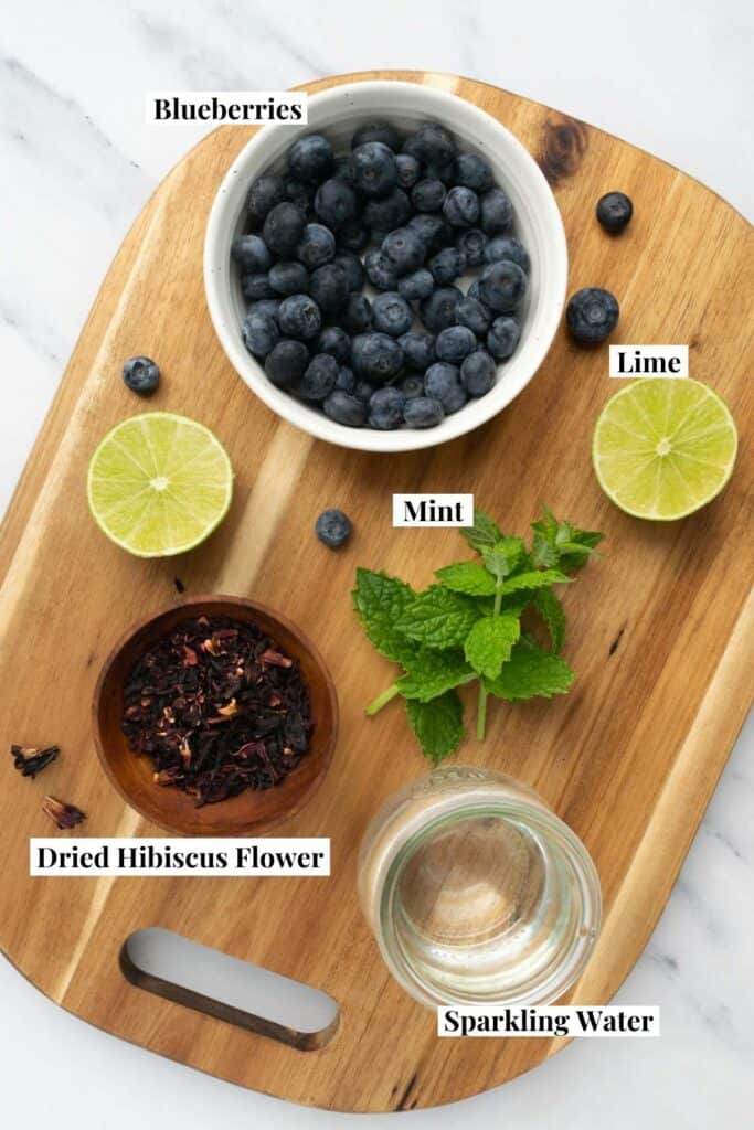 blueberries, lime, dried hibiscus, mint and a jar of sparkling water on a wooden board.