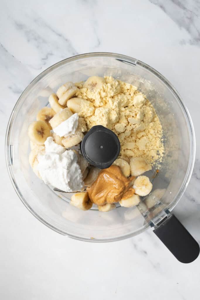 frozen banana, protein powder, peanut butter and coconut cream in the bowl of a food processor.