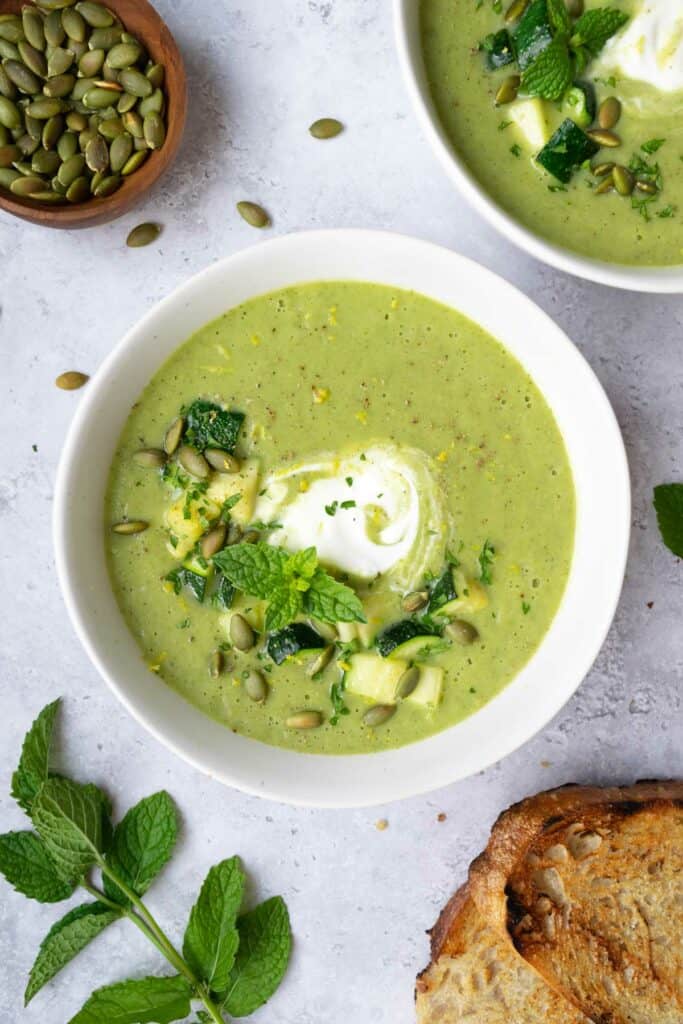 two bowls of zucchini leek soup, a small bowl of pumpkin seeds, toasted bread and a sprig of fresh mint.