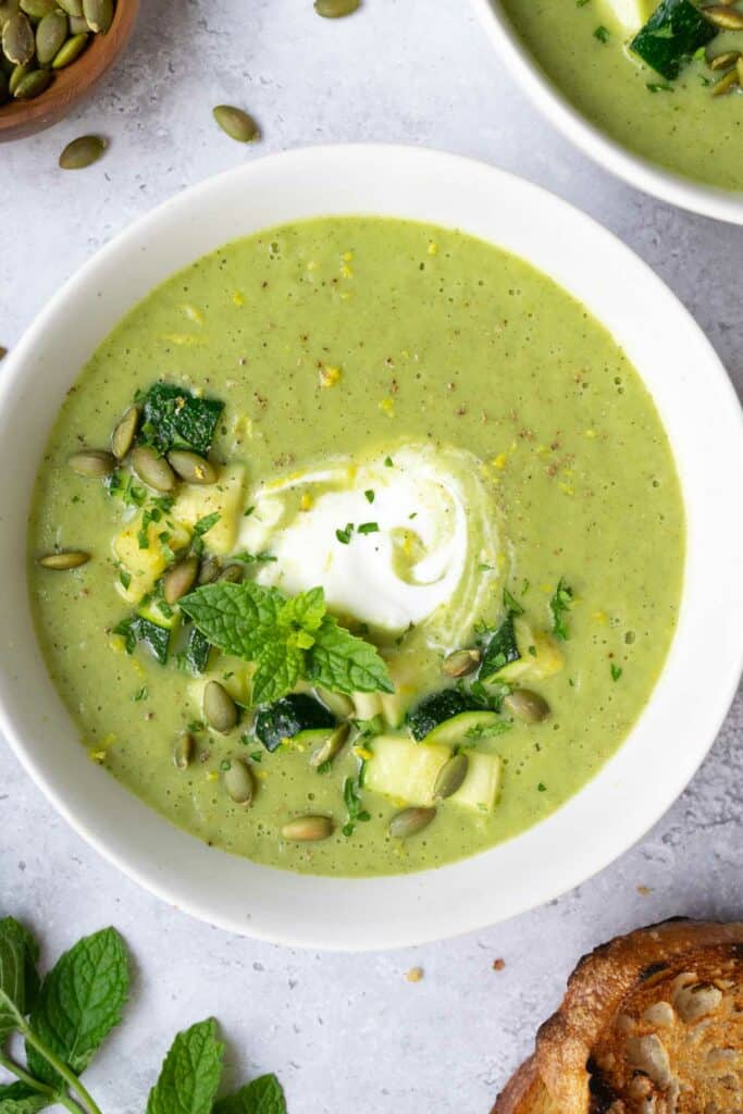 zucchini leek soup in a white bowl topped with yogurt, sautéed zucchini, fresh mint, and toasted pumpkin seeds.