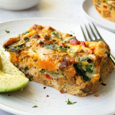 a slice of breakfast casserole on a white plate with avocado, a bowl of oranges and a white mug.