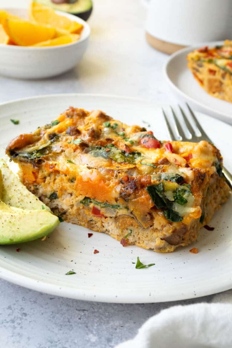 a slice of breakfast casserole on a white plate with avocado, a bowl of oranges and a white mug.