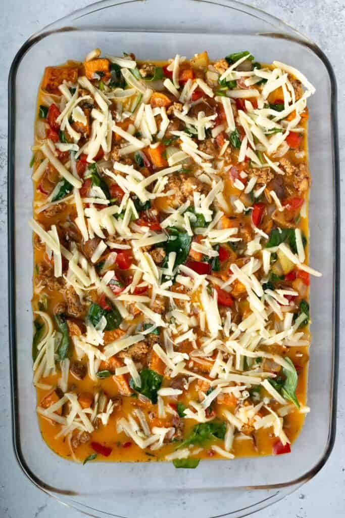 ground meat, roasted sweet potato, cooked vegetables and whisked eggs topped with shredded cheese in a glass casserole dish