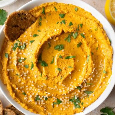 roasted carrot dip in a white bowl, crackers, lemon, and small bowls with hemp seeds and sesame seeds