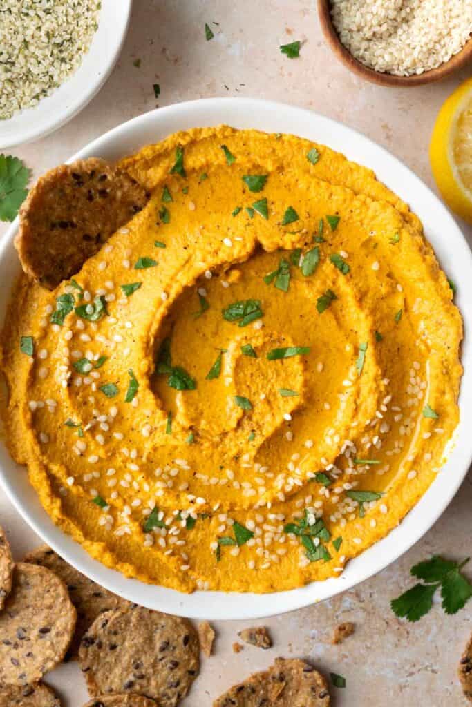 roasted carrot dip in a white bowl, crackers, lemon, and small bowls with hemp seeds and sesame seeds